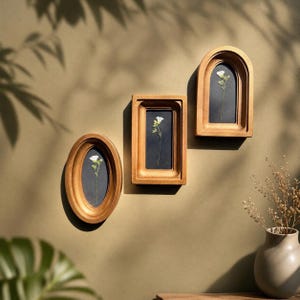 May include: Three wooden framed wall art pieces. Each frame is a different shape, oval, rectangular, and arched. Each frame contains a white flower on a dark background. The frames are a warm gold color and are mounted on a neutral wall.