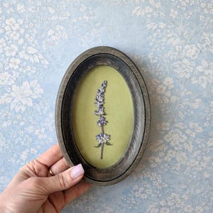 Op de afbeelding: Een ovaal kunstwerk met houten lijst met een takje lavendel op een lichtgroene achtergrond. De lijst is donkergrijs en de achtergrond is een lichtblauw bloemenpatroon.