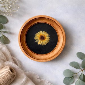 K&ouml;nnte beinhalten: Eine gepresste gelbe Blume mit braunem Zentrum ist in einem runden Holzrahmen zentriert. Die Blume befindet sich vor einem schwarzen Hintergrund. Der Rahmen liegt auf einer wei&szlig;en Oberfl&auml;che mit Gr&uuml;n und einer Schnurrolle.
