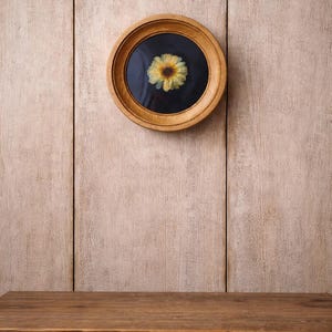 K&ouml;nnte beinhalten: Ein rundes, holzgerahmtes Kunstwerk mit einer gepressten gelben Blume vor einem schwarzen Hintergrund. Der hellbraune Rahmen h&auml;ngt an einer hellbraunen Holzwand. Ein Holztisch befindet sich im Vordergrund.