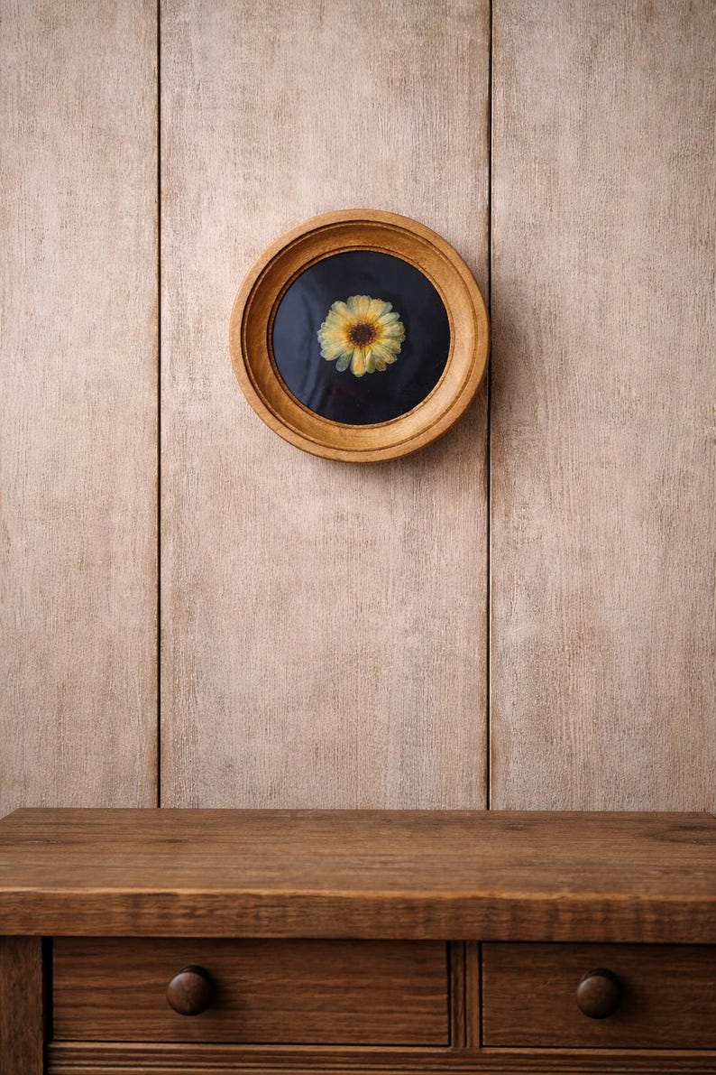 K&ouml;nnte beinhalten: Ein rundes, holzgerahmtes Kunstwerk mit einer gepressten gelben Blume vor einem schwarzen Hintergrund. Der hellbraune Rahmen h&auml;ngt an einer hellbraunen Holzwand. Ein Holztisch befindet sich im Vordergrund.