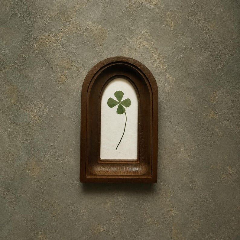 May include: A dark brown wooden frame with an arched top opening containing a single pressed green four-leaf clover centered on a cream-colored background. The frame features a deep, recessed design with visible wood grain and smooth edges.