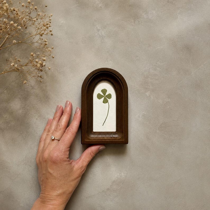 May include: A dark brown wooden arch-shaped frame containing a white background with a single green four-leaf clover illustration centered inside. The frame has a smooth, rounded top edge and a flat base, presenting a clean and simple display.