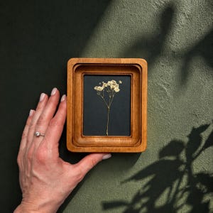 Könnte beinhalten: Ein kleiner, quadratischer Holzrahmen mit dunklem Hintergrund, der eine zarte Trockenblume zeigt. Der Rahmen hat ein mehrschichtiges Design und einen warmen, natürlichen Holzton. Die Blume hat eine hellbeige Farbe und verleiht dem Stück einen Hauch von Natur.