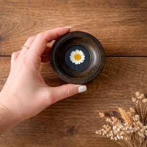 May include: A small, dark wooden bowl with a pressed daisy flower encased in a dark blue background. The daisy has white petals and a yellow centre. The bowl is held against a wooden surface.