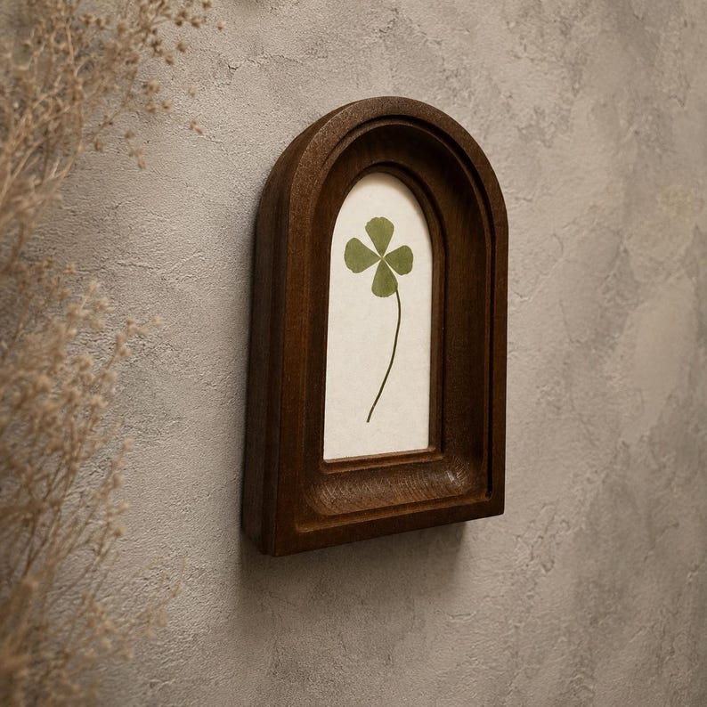 May include: A dark brown wooden wall frame with an arched top design. The frame contains a light-colored backing featuring a single pressed green four-leaf clover centered within the display area. The frame has a deep, recessed border.