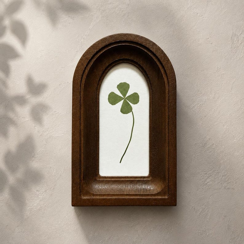 May include: A dark brown wooden frame with an arched top containing a white background. Centered on the white background is a single pressed green four-leaf clover with a thin stem. The frame has a deep, recessed design with smooth edges.