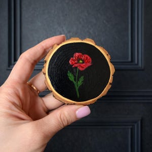 以下が含まれることがあります： 丸い木の輪切りに手描きされた赤いポピーの花。ポピーは緑色の茎と葉を持ち、黒い背景に配置されています。木の輪切りは自然な樹皮の縁があり、素朴な美しさを作り出しています。
