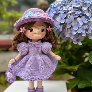 Puede incluir: Muñeca de ganchillo hecha a mano con vestido, sombrero y zapatos morados. La muñeca tiene cabello castaño, detalles rosas y un pequeño bolso. Un arbusto de hortensias azules en el fondo.
