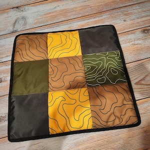 Può includere: Cuscino quadrato con un design patchwork. Il cuscino presenta nove quadrati nei toni del giallo, marrone, verde oliva e grigio. Ogni quadrato ha un design a mappa topografica in nero. Il cuscino è rifinito con un bordo nero.