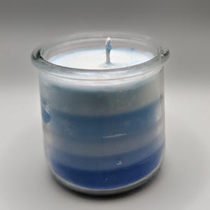 May include: A blue and white layered candle in a clear glass jar. The candle has a wick in the center and is made with different shades of blue and white. The jar has a thick rim.