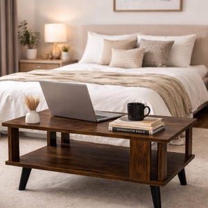 May include: A dark brown rectangular coffee table with a lower shelf, black legs, and a laptop. A black mug and books are on the table. A small white vase with dried grass sits on the table. The table is in front of a bed.
