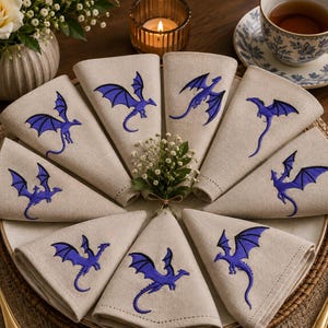 May include: A set of eight beige linen napkins arranged in a circular pattern on a plate, each featuring a single embroidered purple dragon. The napkins are folded into triangles, showcasing the detailed stitching of the mythical creatures.