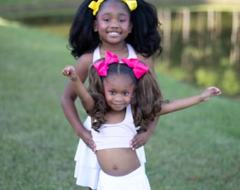 May include: Two young girls in white outfits with large bows in their hair. One has yellow bows, the other pink. They are standing on grass near a pond or lake.