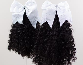 May include: Two cheerleading hair bows. Each bow has a white satin bow with silver rhinestone detailing and a cluster of black, curly hair. The bows are set against a white backdrop.