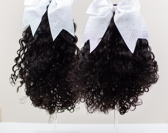 May include: Two cheerleading hair bows with black curly hair extensions. Each bow is white with a rhinestone design and a small rhinestone accent in the centre. The hair extensions are attached to clear stands.
