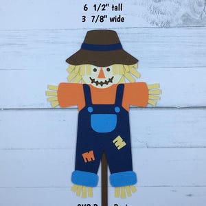 May include: A handmade paper scarecrow cutout, 6.5 inches tall and 3.875 inches wide. It features a brown hat, straw hair, an orange shirt, blue overalls with yellow patches, and straw feet. The text "CKS Paper Designs Handmade Die Cuts" is at the bottom.