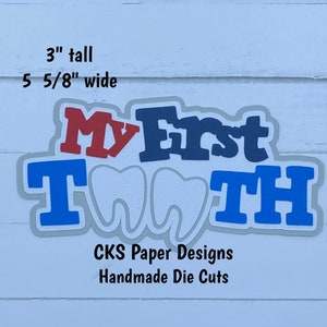 May include: A handmade die cut of the words "My First Tooth" in blue, red, and gray letters with two white teeth in the center. The die cut is 3 inches tall and 5 5/8 inches wide.