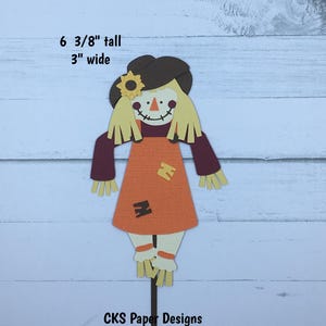 May include: A papercraft scarecrow die cut, 6 3/8 inches tall and 3 inches wide. The scarecrow is wearing a brown hat with a yellow sunflower, an orange dress with brown patches, and brown sleeves. The scarecrow has a yellow face with a stitched smile and a brown nose.