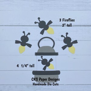 May include: Three black paper fireflies with yellow accents, each measuring 2 inches tall, and a black paper lantern with a yellow firefly inside, measuring 4 1/4 inches tall. The text "3 Fireflies 2" tall" and "4 1/4" tall" is printed on the white background. The text "CKS Paper Designs Handmade Die Cuts" is printed below the lantern.