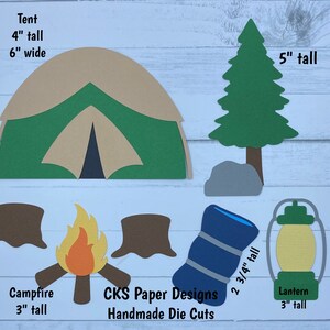 May include: A set of paper die cuts for a camping theme. The set includes a tent, a tree, a campfire, a lantern, and a sleeping bag. The tent is 4 inches tall and 6 inches wide. The tree is 5 inches tall. The campfire is 3 inches tall. The lantern is 3 inches tall. The sleeping bag is 2 3/4 inches tall. The die cuts are made by CKS Paper Designs.
