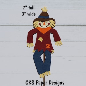 May include: A 7-inch tall, 3-inch wide paper die cut of a scarecrow with a brown hat, red shirt, blue pants, and a straw-like beard. The scarecrow is standing on a stick. The text "CKS Paper Designs Handmade Die Cuts" is at the bottom of the image.