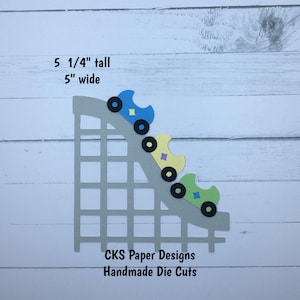 May include: A paper die-cut of a roller coaster with three colorful cars. The cars are blue, yellow, and green, with black wheels. The roller coaster is 5 1/4 inches tall and 5 inches wide. The text "CKS Paper Designs Handmade Die Cuts" is at the bottom.