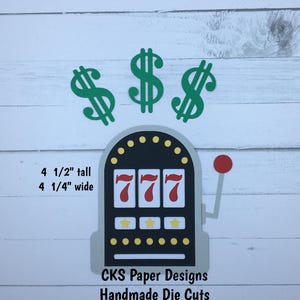 May include: A die cut of a slot machine with three red sevens, three yellow stars, and a red lever. Three green dollar signs are above the slot machine. The slot machine is 4 1/2 inches tall and 4 1/4 inches wide. CKS Paper Designs Handmade Die Cuts.
