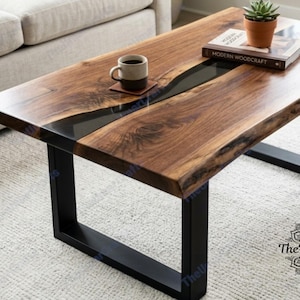 Puede incluir: Una mesa de centro rectangular con una tapa de madera de borde vivo y una incrustación de resina epoxi negra. La mesa tiene patas de metal negro y un acabado de grano de madera natural. Una taza de café, un posavasos, un libro y una pequeña planta en maceta están sobre la mesa.