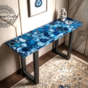 May include: A rectangular console table featuring a polished blue agate stone slab top with varied shades of azure and navy, supported by two dark grey metal U-shaped legs. The surface displays a mosaic of natural stone patterns and textures.