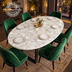May include: An oval dining table with a white, textured stone surface and a black metal base, surrounded by six emerald green velvet chairs with gold-toned accents. The table is set with white plates, a glass vase with flowers, and two small candles.