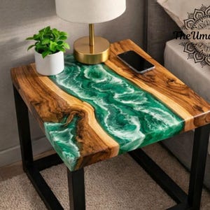Puede incluir: Una mesa auxiliar cuadrada con una tapa de madera y un diseño de río de resina epoxi verde y blanca. La mesa tiene una estructura de metal negro. Una pequeña planta en maceta, una lámpara y un teléfono inteligente están sobre la mesa.