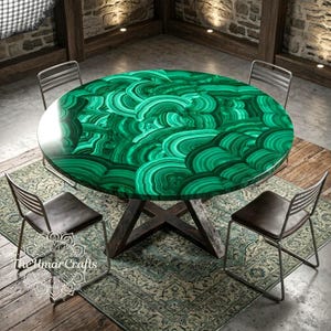 Può includere: Un tavolo da pranzo rotondo con una superficie dal motivo in malachite verde brillante con bande concentriche vorticose. Il tavolo è sostenuto da una base in metallo scuro a forma di X ed è circondato da quattro sedie in metallo con sedute marrone scuro. Il testo The Umar Crafts è visibile.
