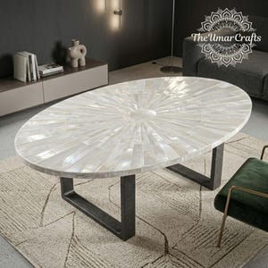 Könnte beinhalten: Ovaler Couchtisch mit einer weißen, perlmutt-eingelegten Platte. Der Tisch hat ein Sonnenstrahlenmuster und steht auf dunkelgrauen Metallbeinen. Das Umar Crafts-Logo ist oben rechts sichtbar.