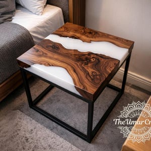 Könnte beinhalten: Quadratischer Beistelltisch mit einer Holzplatte mit einem Fluss aus weißem Harz. Das Holz hat eine satte, dunkelbraune Maserung. Der Tisch hat ein schwarzes Metallgestell. Das Umar Crafts-Logo befindet sich unten rechts.