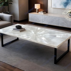 Puede incluir: Mesa de centro rectangular blanca con un patrón radial en la superficie. La mesa tiene patas de metal negro y está sobre una alfombra gris. Un cuenco negro y libros están sobre la mesa.
