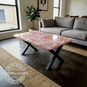 May include: A rectangular coffee table featuring a polished, translucent pink rose quartz slab top with visible natural crystalline fractures. The tabletop is supported by a black metal base with two intersecting X-shaped legs.