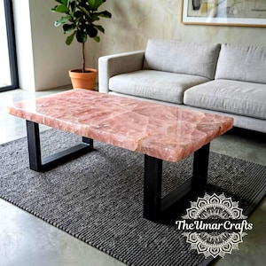 May include: A rectangular coffee table featuring a polished, translucent pink rose quartz slab top with natural crystalline textures, supported by two sturdy, matte black metal U-shaped legs. The surface displays a smooth, reflective finish.