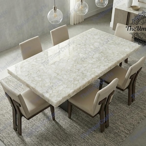 May include: A rectangular dining table with a white, patterned surface, surrounded by six beige chairs with dark wooden legs. The table sits on a grey rug, and the room has pendant lights.