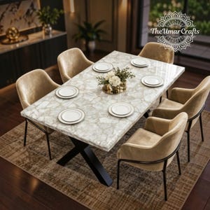 Può includere: Un tavolo da pranzo rettangolare con piano a motivo pietra bianca e base in metallo nero a forma di X, circondato da sei poltrone imbottite beige. Ogni posto tavola presenta un piatto bianco. Il logo The Umar Crafts è visibile.