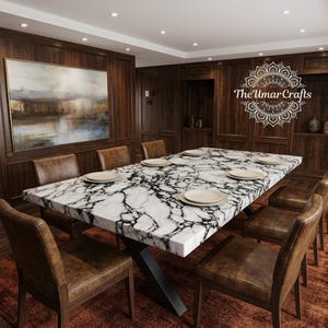 Puede incluir: Una mesa de comedor rectangular con tablero de mármol blanco con vetas negras y base de metal negro, rodeada de sillas de cuero marrón. La mesa está puesta con platos blancos. El logotipo The Umar Crafts es visible en la pared de madera.