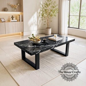 May include: A rectangular coffee table featuring a polished black marble top with white veining, supported by two sturdy black metal U-shaped legs. The table surface holds a small bowl of dried flowers, a black coffee cup, and a book.