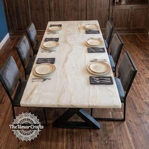 Op de afbeelding: Een rechthoekige eettafel met een wit, doorschijnend stenen blad met subtiele beige aders en een zwart metalen X-vormig onderstel. De tafel is gedekt met zes grijze gestoffeerde stoelen, witte borden en zilveren bestek op donkere servetten.