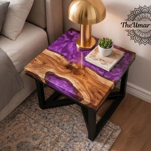 Puede incluir: Una mesita de noche artesanal con un diseño único. La mesa presenta una tapa de madera con una incrustación de resina púrpura, sostenida por un marco de metal negro. Una lámpara dorada, una pequeña planta y un libro con gafas descansan sobre la mesa. El logotipo de Umar Crafts es visible.