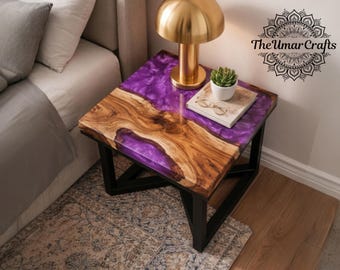May include: A handcrafted side table with a unique design. The table features a wooden top with a purple resin inlay, supported by a black metal frame. A gold lamp, a small plant, and a book with glasses rest on the table. The Umar Crafts logo is visible.
