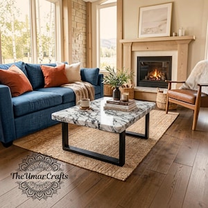 Pode incluir: Uma mesa de centro retangular com um tampo de padrão de mármore branco e preto e uma estrutura de metal preta. A mesa está posicionada sobre um tapete tecido numa sala de estar, com um sofá azul com almofadas laranja e creme e uma poltrona de couro.