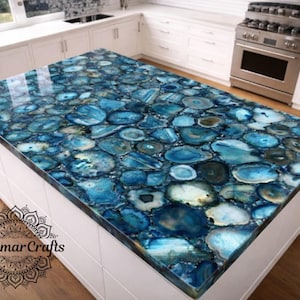 May include: A kitchen island with a countertop made of blue agate stone. The stone is composed of various shades of blue and white, creating a unique pattern. The island has a white base and is set in a modern kitchen with white cabinets and stainless steel appliances.