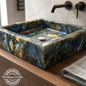 May include: A square, stone bathroom sink with a unique, iridescent pattern of blues, greens, and golds. The sink has a polished chrome drain and is set on a wooden countertop. A black faucet and towel are also visible.