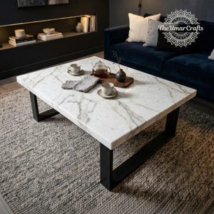 May include: A rectangular coffee table with a white marble top featuring grey veining and a black metal frame. On the table sits a glass teapot, two white ceramic cups on saucers, and a grey cloth napkin on a wooden tray. The table rests on a rug.