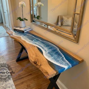 May include: A wooden console table with a blue and white resin river design. The table has a natural wood grain finish and black metal legs. The resin mimics the look of ocean waves. The table is placed against a wall with a mirror.